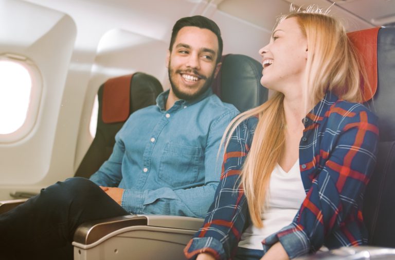 On a Commercial Plane Flight Handsome Hispanic Man Tells Funny Story to His Beautiful Blonde Girlfriend. Both Laugh. They Travel in New Airplane, with Sun Shining Through the Window.